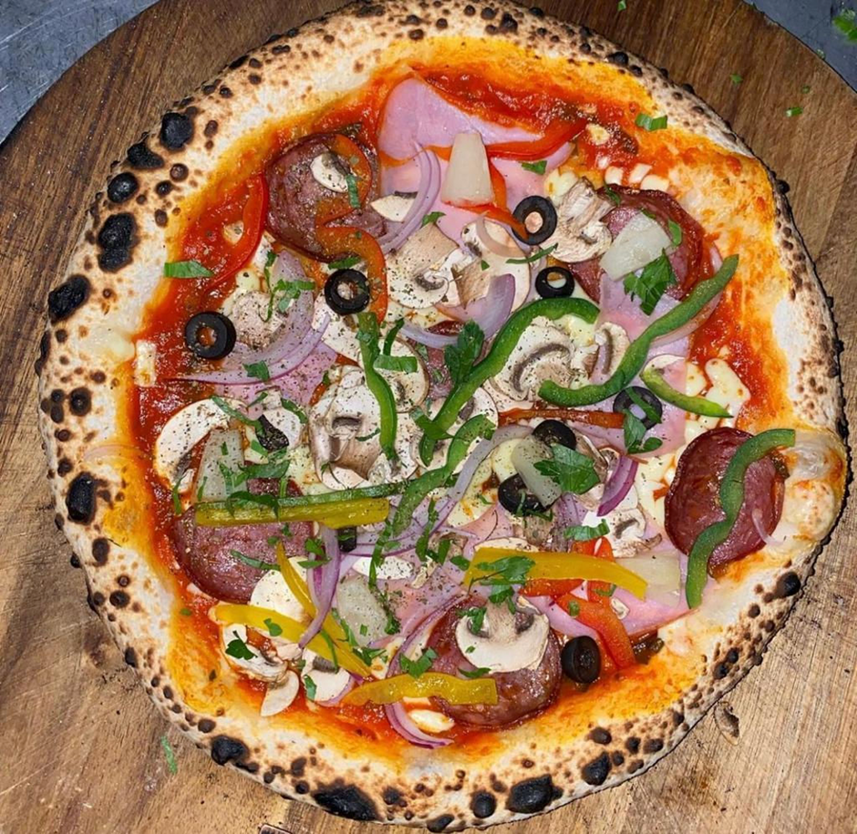 Top-down view of a loaded wood-fired pizza with capsicum, mushroom, olives and cured meat